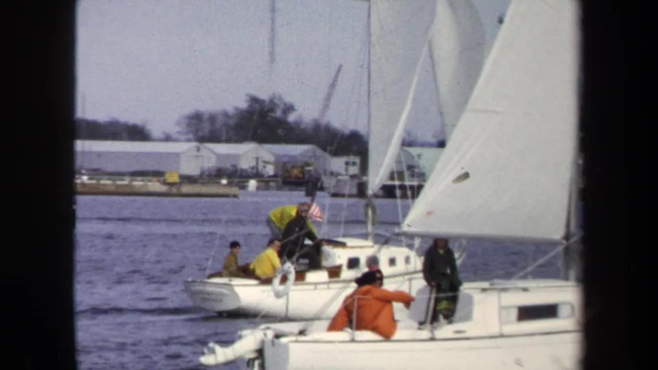 1970 Annapolis home-movie frame of white sailboats with people aboard, seen from another boat near a low waterfront shoreline.