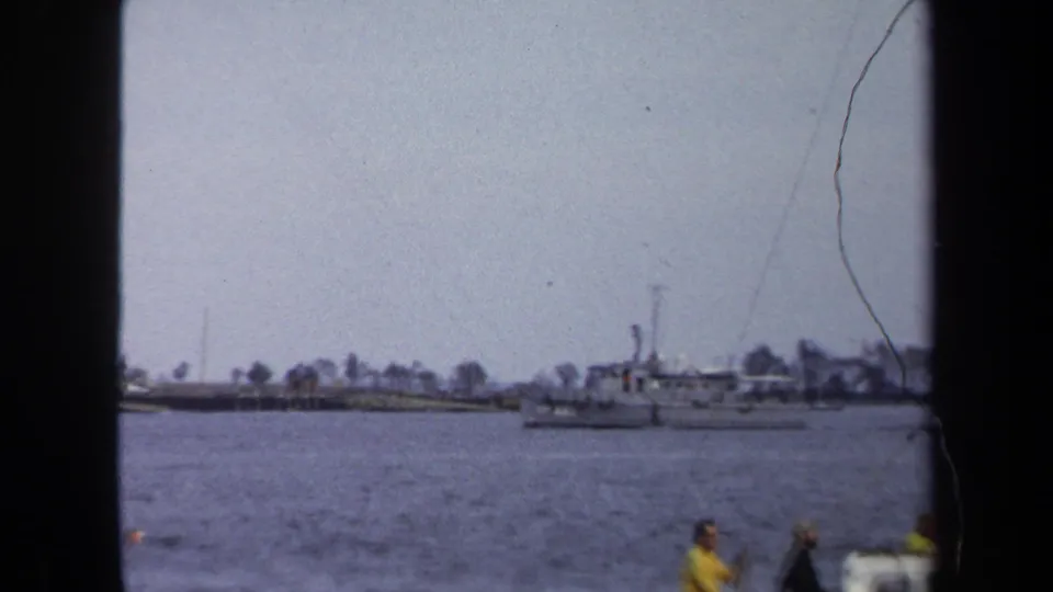 1970 Annapolis home-movie frame showing a distant motor vessel on bay water, shoreline trees, and partial people near the camera.