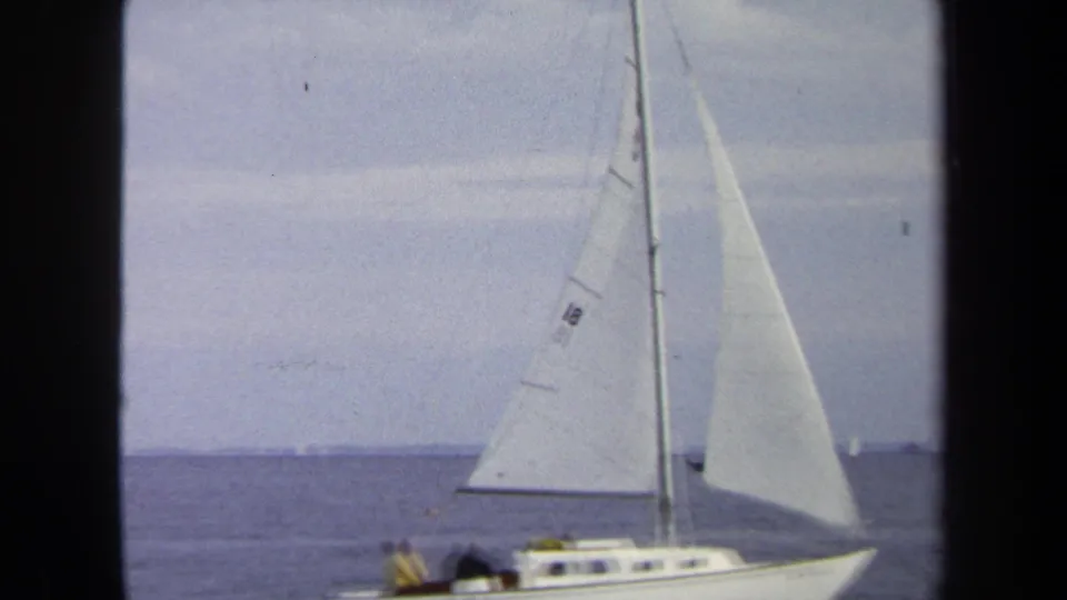 1970 Annapolis frame showing a white sailboat with raised sails moving across open bay water under a pale sky.