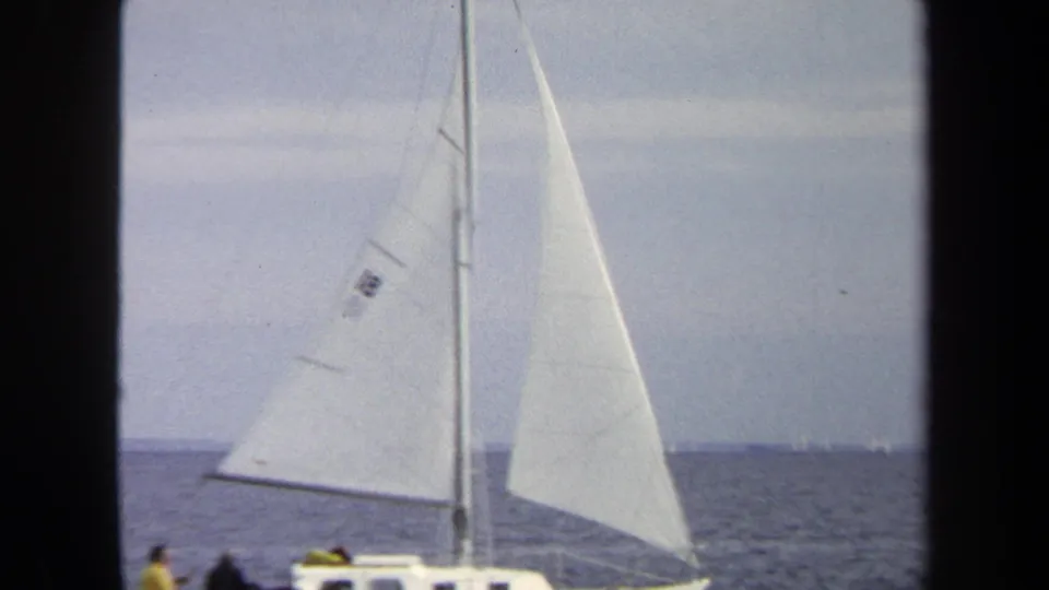 1970 Annapolis home-movie frame showing a nearby sailboat with white sails and small figures on open water.