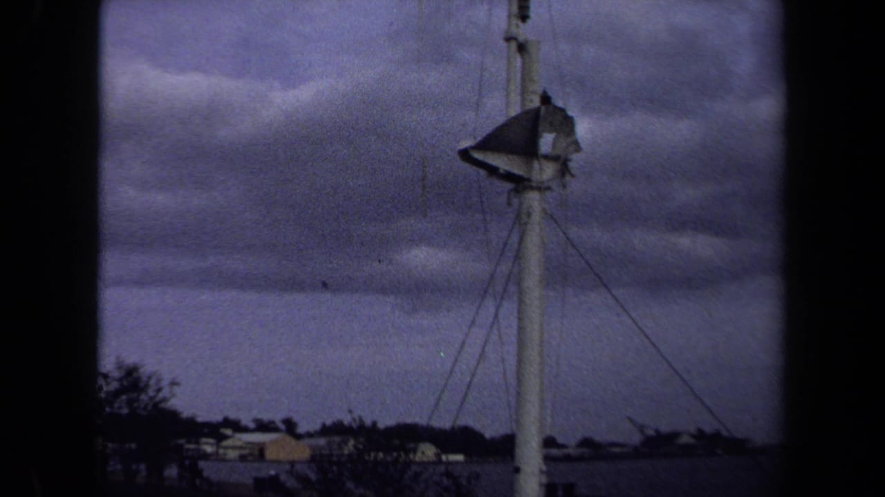 1970 8mm Annapolis footage showing a tall white pole or tower with a weathered flag below a cloudy sky and low shoreline buildings.