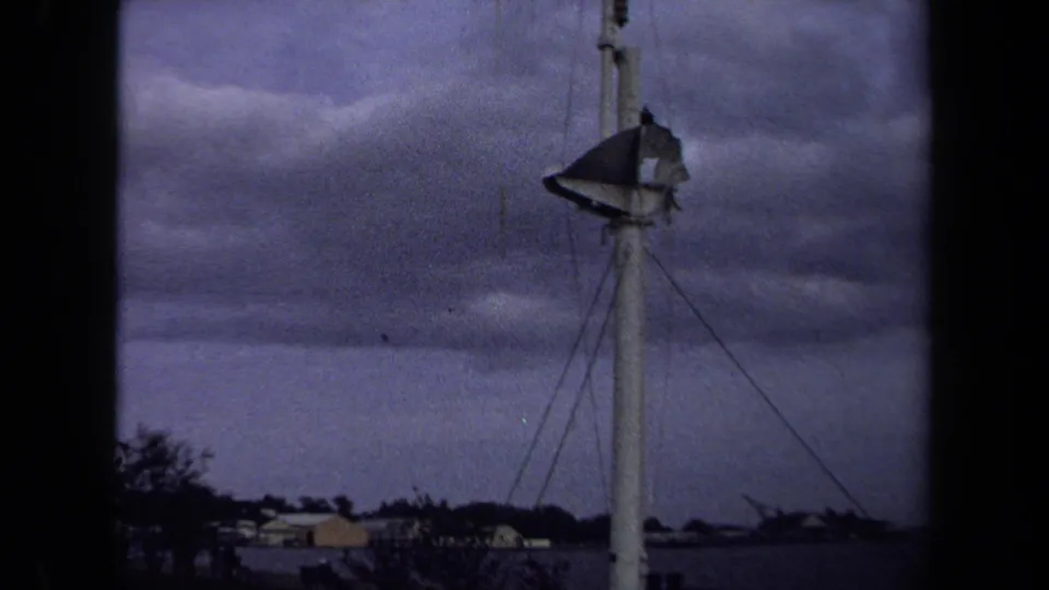1970 8mm Annapolis footage showing a tall white pole or tower with a weathered flag below a cloudy sky and low shoreline buildings.