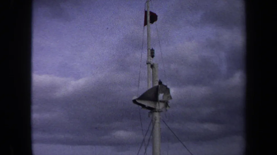 1970 8mm frame of the tower rising into heavy clouds, with a small flag near the top and a larger tattered flag lower on the structure.