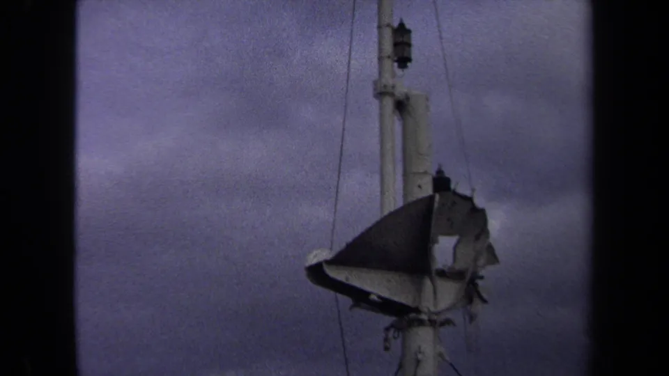 Close 8mm view of the tattered flag mounted on the white tower, with wires and a cloudy Annapolis sky filling the background.