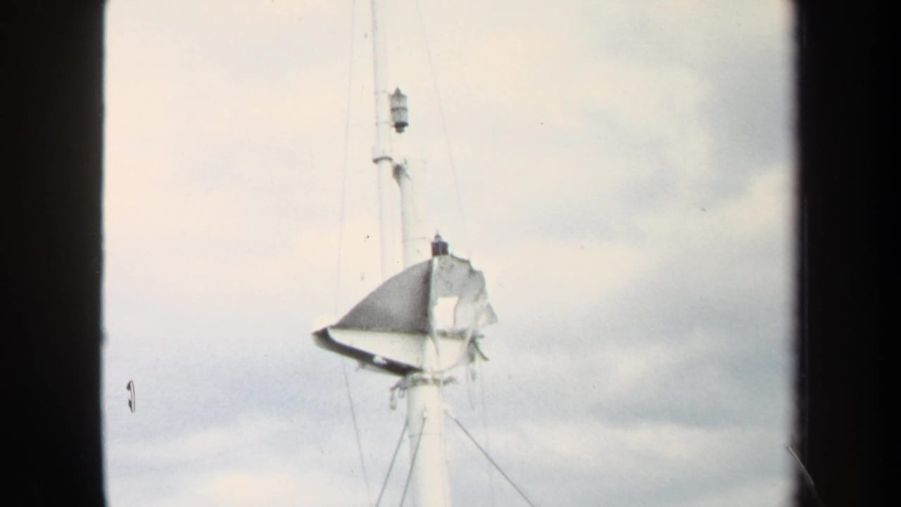 White mast with torn dark material and rigging centered against pale clouds.