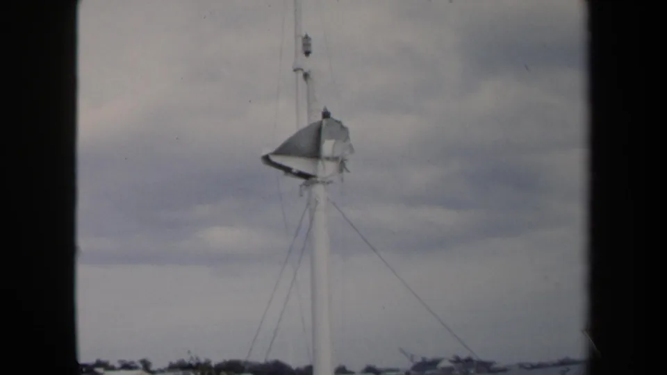 1970 Annapolis frame showing the damaged mast, slack rigging, cloudy sky, and a low shoreline in the distance.