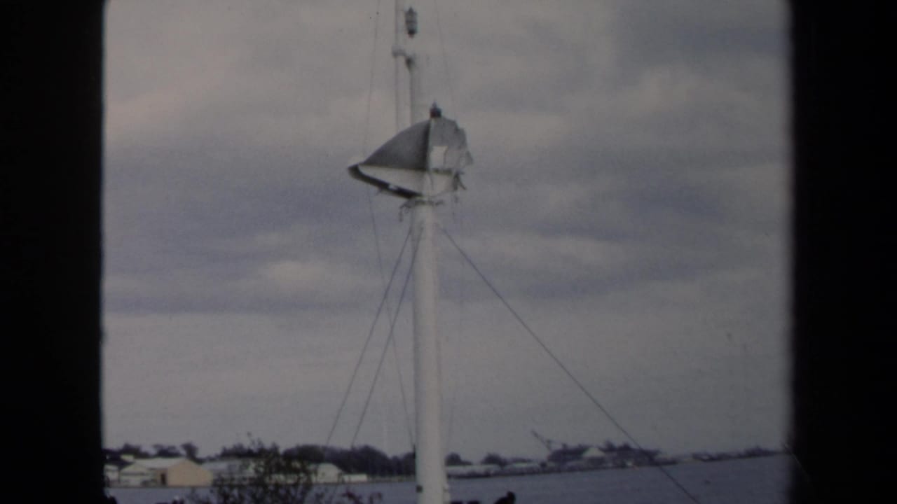 1970 Annapolis 8mm frame showing a sailboat mast, rigging lines, cloudy sky, distant shoreline, and dark film-frame edges.