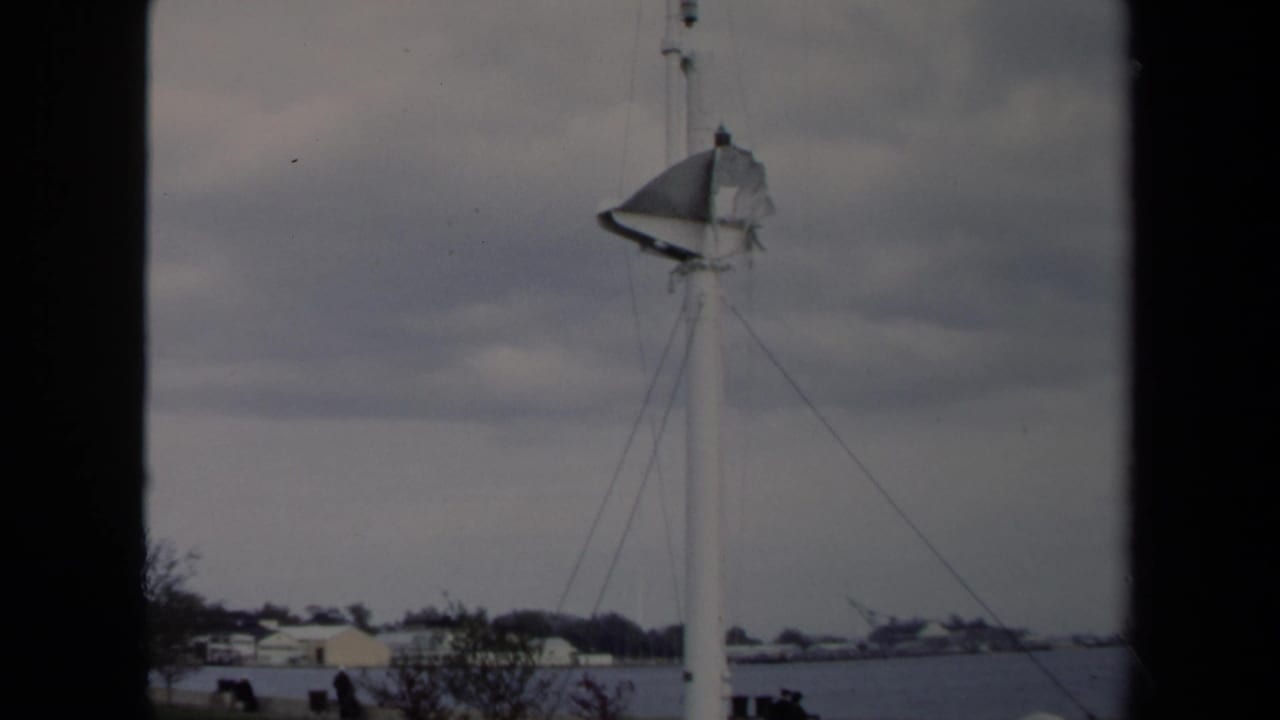 Sailboat mast beside a cloudy waterfront with low buildings and dark frame borders.