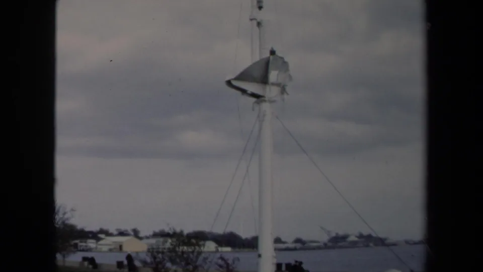 1970 Annapolis view of a sailboat mast and rigging in front of an overcast sky, with shoreline buildings and small distant figures.