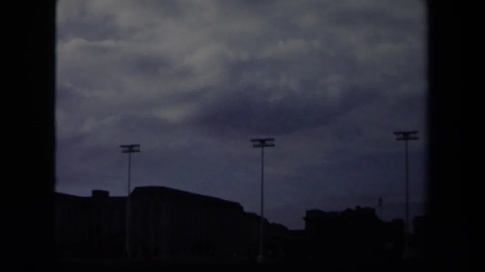 1970 Annapolis 8mm frame with tall stadium light poles, dark foreground buildings, overcast clouds, and strong film-edge shadowing.