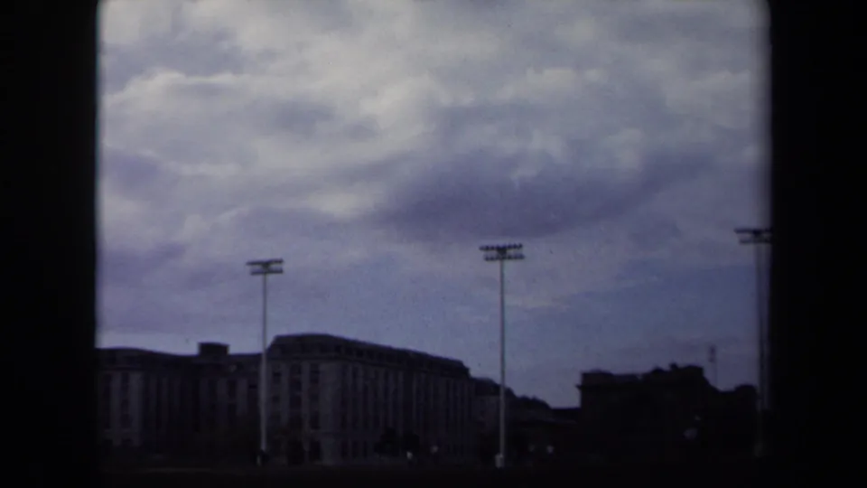 1970 Annapolis frame showing stadium light poles, a large distant building, dark lower skyline, and textured cloudy sky on 8mm film.