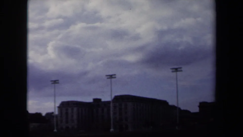 1970 Annapolis frame with tall sports lights and a broad stadium building silhouetted against an overcast daytime sky.