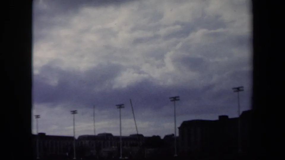 1970 Annapolis frame showing multiple stadium light poles, distant buildings, and a dramatic overcast sky on 8mm film.