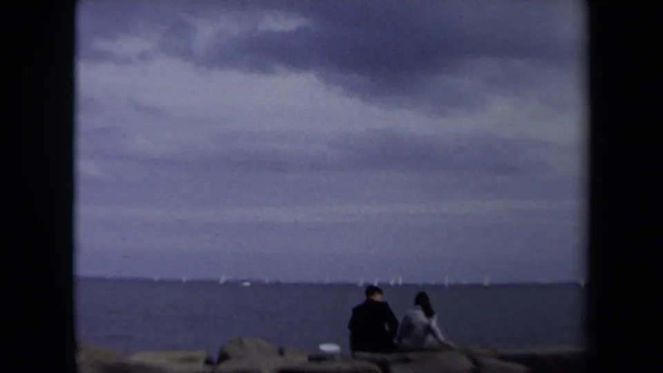 Two distant figures sit on a rocky Annapolis shoreline facing open water and faint sailboats beneath a cloudy sky.