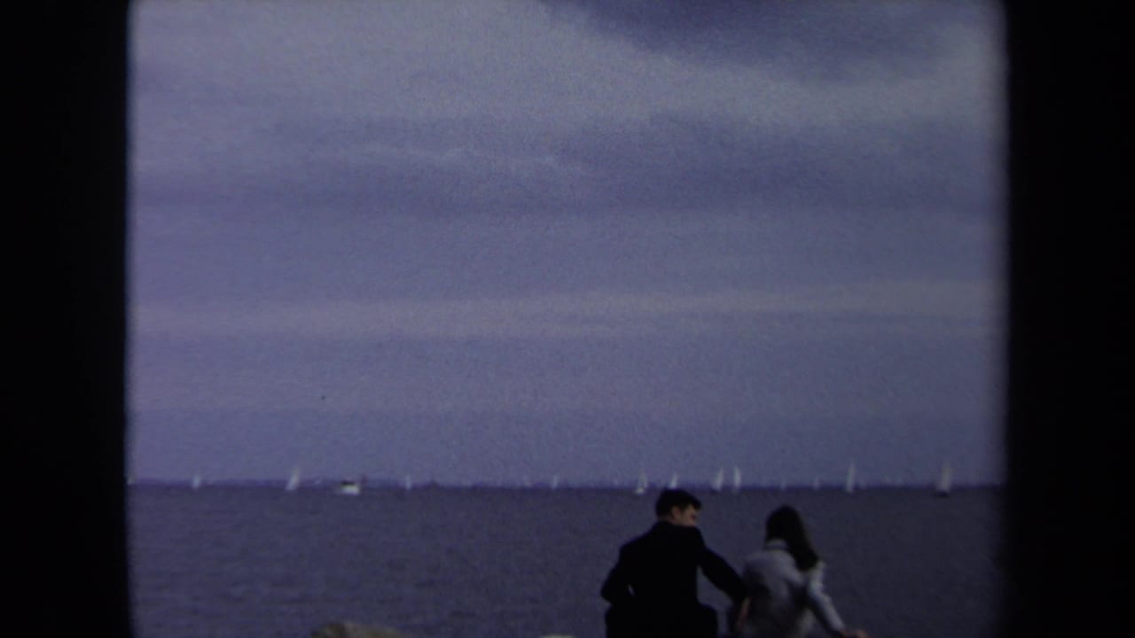 In supported 1970 Annapolis home-movie footage, a couple sits near the water under an overcast sky with small sailboats in the distance.