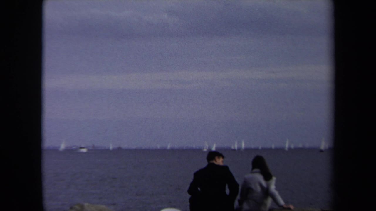 Two adults sit near the water facing distant sailboats beneath a cloudy sky.