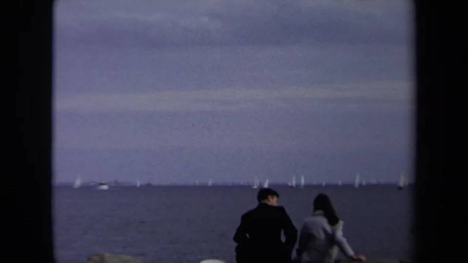 The couple remains seated on the shore in the 1970 Annapolis footage, framed against open water and a line of small sailboats.
