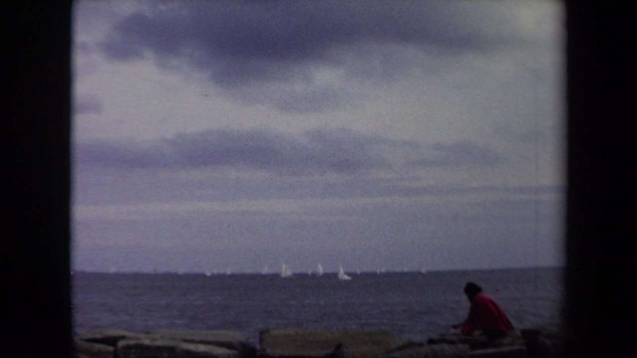 In 1970s Annapolis home-movie footage, two dark-clad figures sit on rocks beside the water as small white sailboats cross the horizon.
