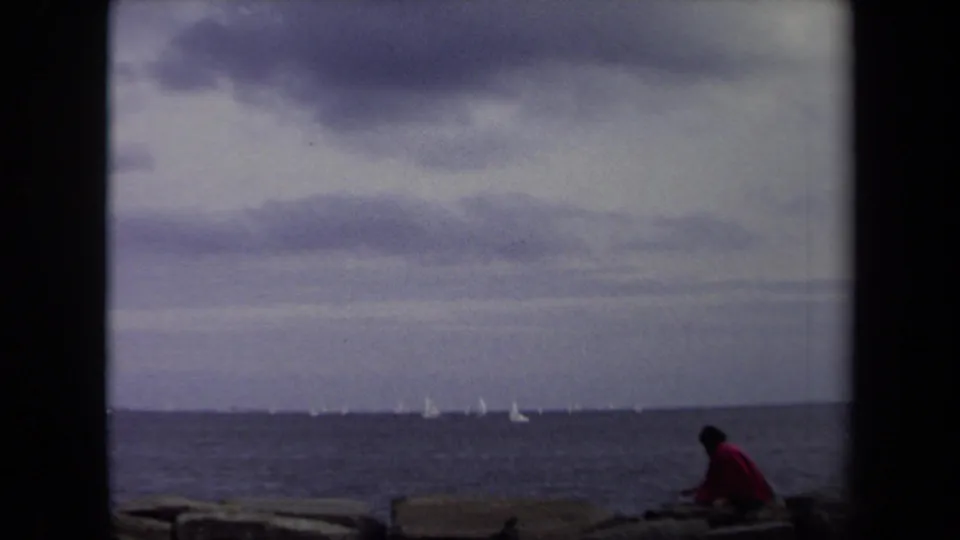 In 1970s Annapolis home-movie footage, two dark-clad figures sit on rocks beside the water as small white sailboats cross the horizon.