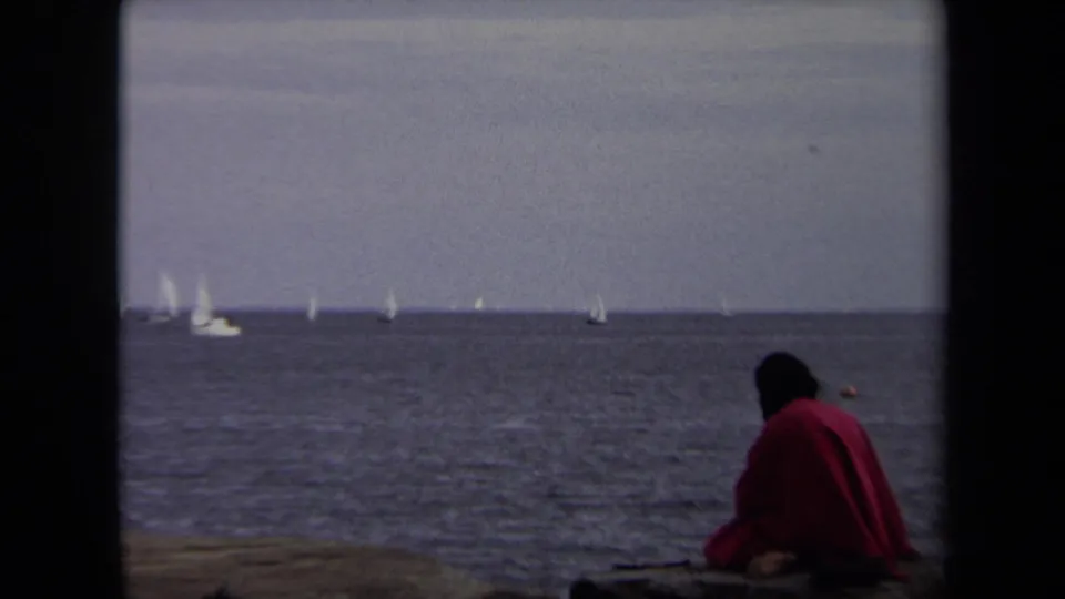 The seated figure in a red jacket is closer in the foreground, facing away toward a wide Annapolis water view with white sails on the horizon.