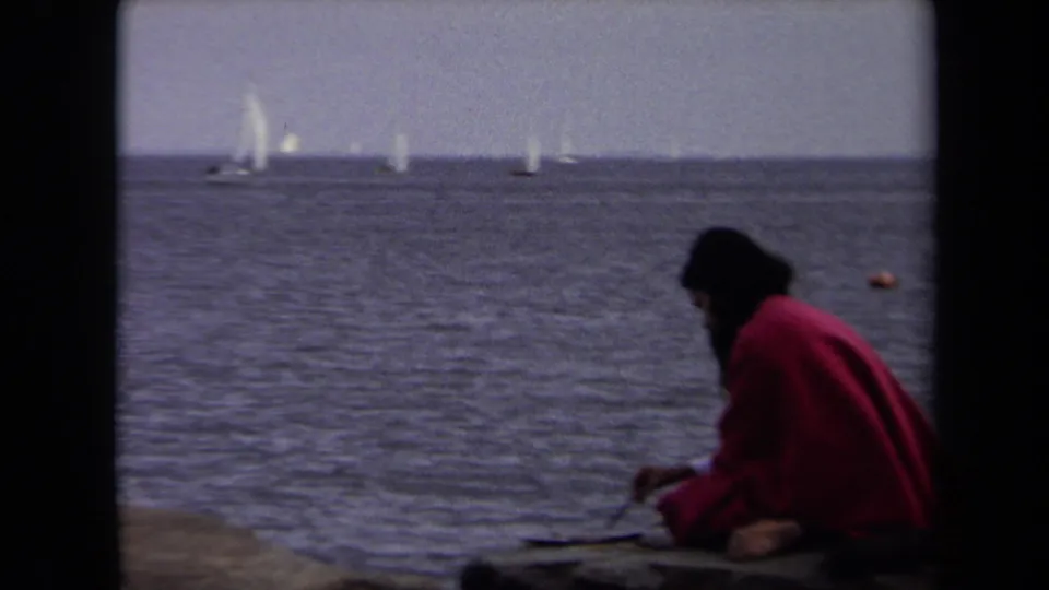 In supported 1970 Annapolis footage, a woman in a red coat sits at the waterfront with sailboats on the horizon.