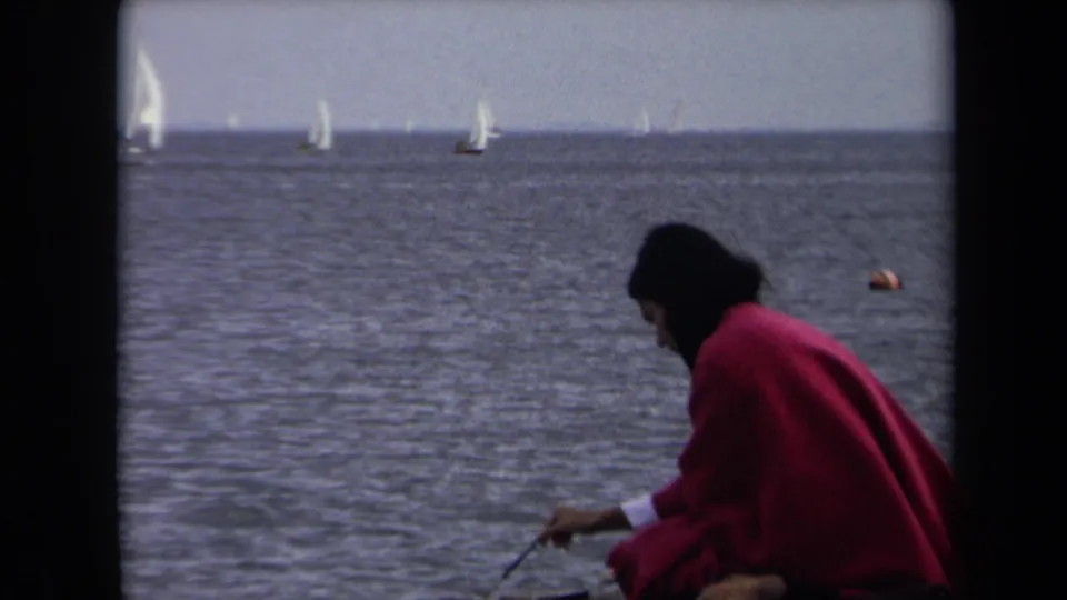 A woman in a red coat sits near the water in Annapolis, with several sailboats visible across the river or bay.