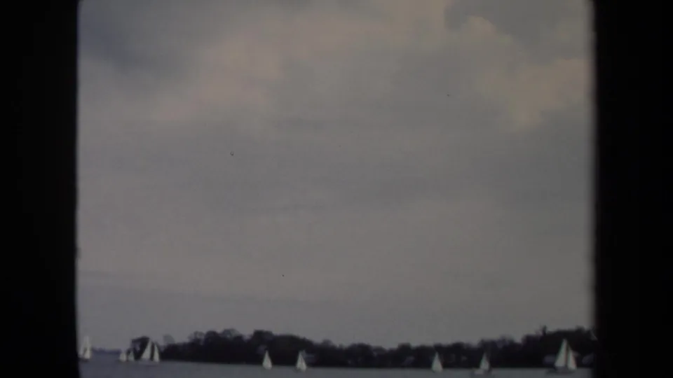 1970 Annapolis home-movie still showing distant sailboats scattered across calm water and a low shoreline.