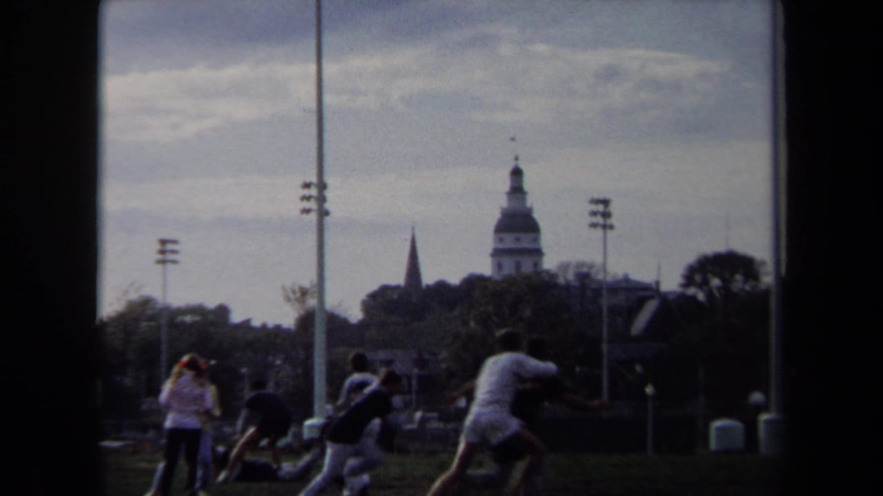1970 Annapolis home-movie frame showing people playing on grass with a domed skyline, spire, and waterfront light poles in the background.