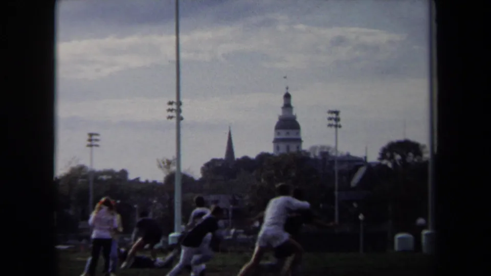 1970 Annapolis home-movie frame showing people playing on grass with a domed skyline, spire, and waterfront light poles in the background.
