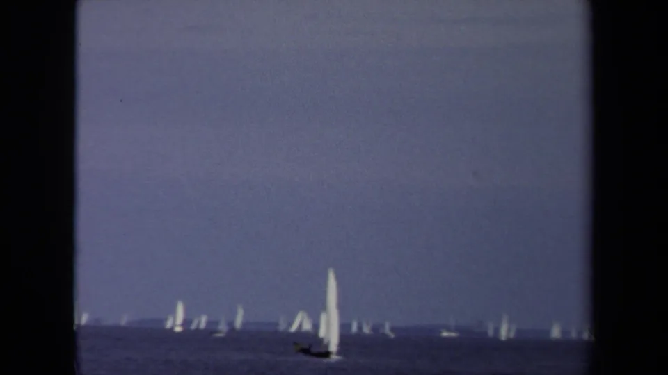 1970 Annapolis home-movie frame of a white-sailed boat crossing calm water with many smaller sails along the horizon.
