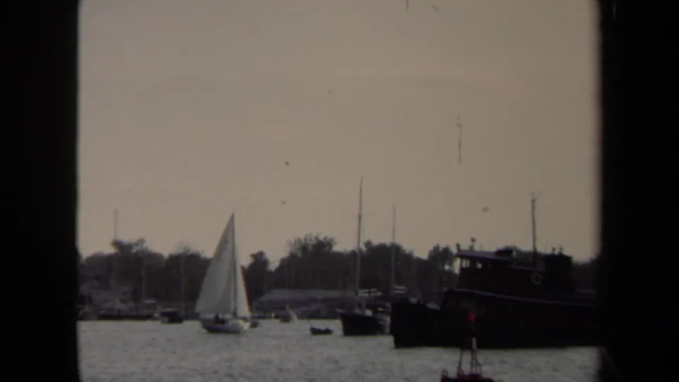 1970 Annapolis harbor frame showing a white sailboat near shore, masts, small boats, trees, and a dark moored vessel at right.