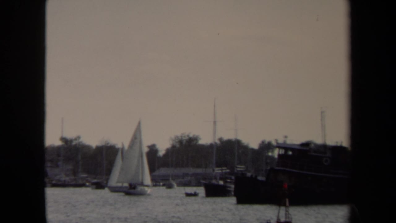 1970 Annapolis harbor view with a white sailboat, rippled water, moored craft, shoreline trees, and dark window-like borders.