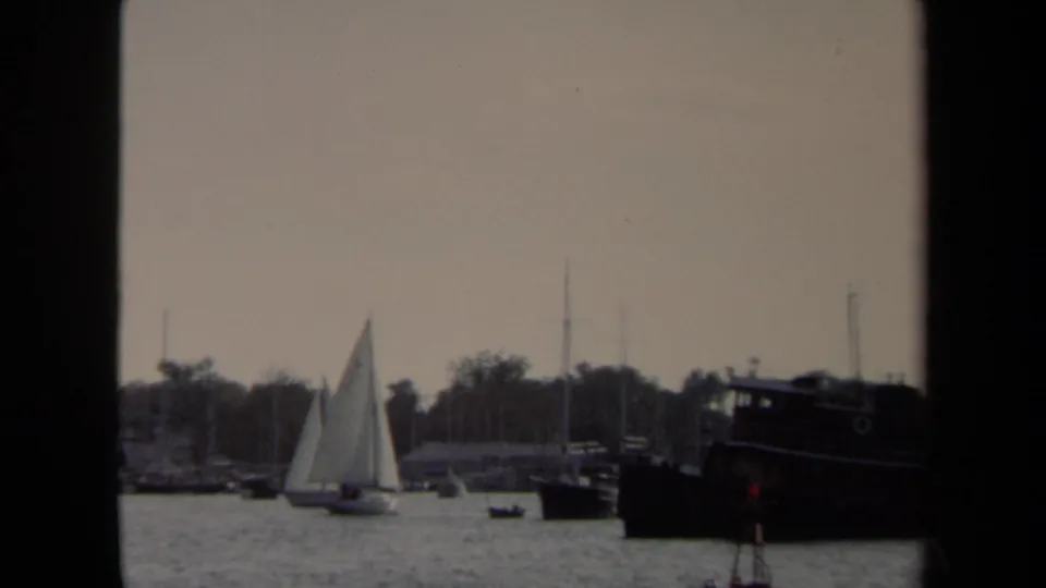 1970 Annapolis harbor view with a white sailboat, rippled water, moored craft, shoreline trees, and dark window-like borders.