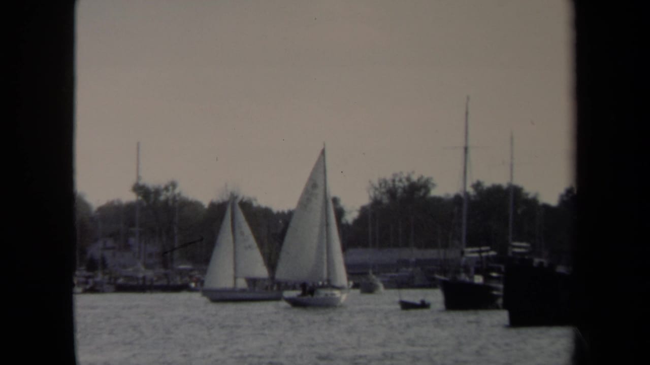 Two white sailboats cross the 1970 Annapolis harbor with background masts, tree-lined shore, and a dark vessel at right.
