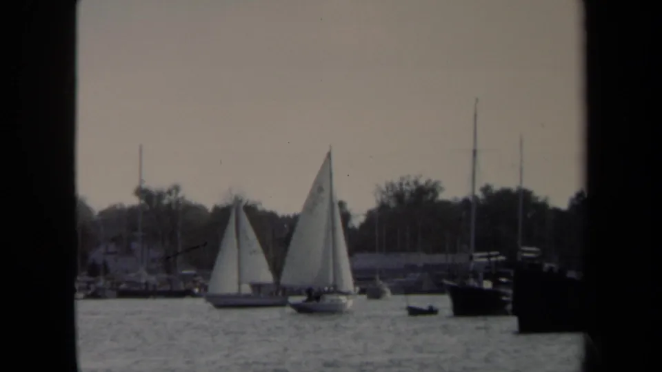 Two white sailboats cross the 1970 Annapolis harbor with background masts, tree-lined shore, and a dark vessel at right.
