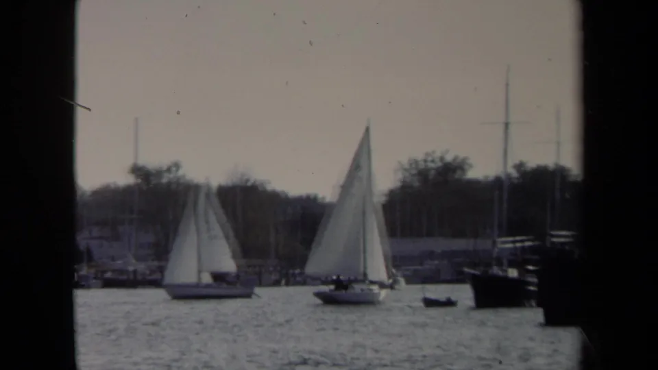 1970 Annapolis harbor scene showing two sailboats under white sails, moored craft, masts, and a soft tree-lined background.