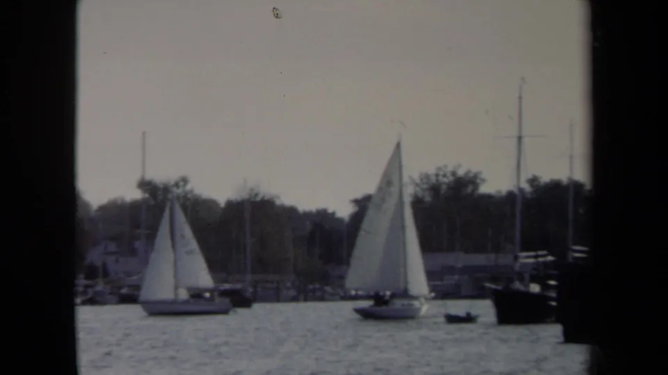 Two white sailboats continue across the Annapolis harbor in 1970, framed by water, small craft, masts, and shoreline trees.