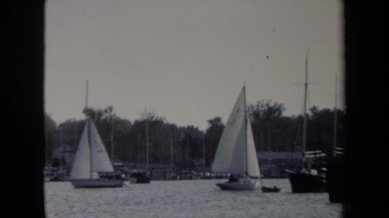 1970 8mm Annapolis waterfront frame with sailboats on rippled water, marina masts, shoreline trees, and dark window-like borders.