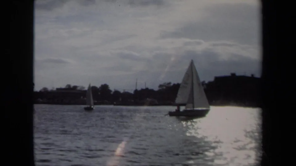 Annapolis waterfront footage shows a sailboat silhouetted against sun glare on rippled water, with buildings and a distant steeple-like shape.