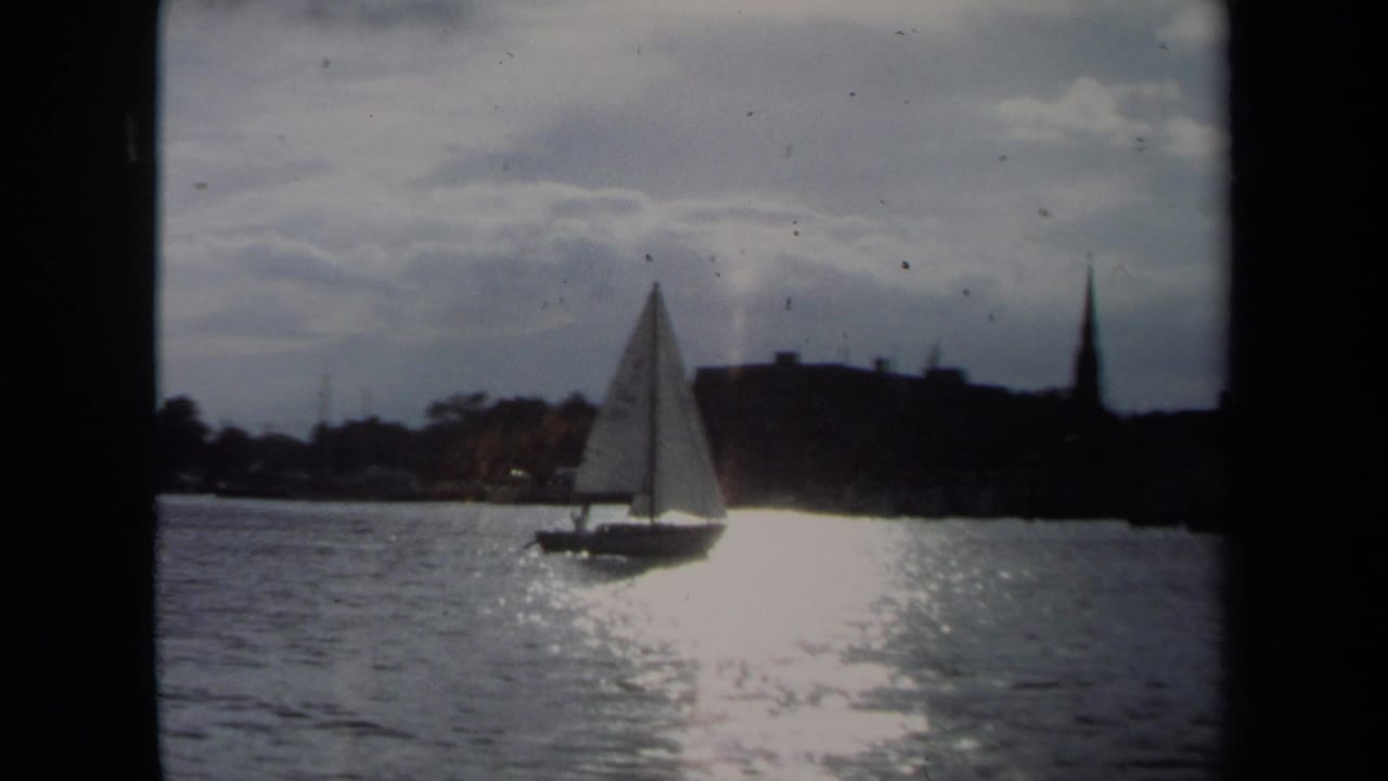 1970 Annapolis harbor view with a white sailboat moving across reflective water and a steeple in the background.