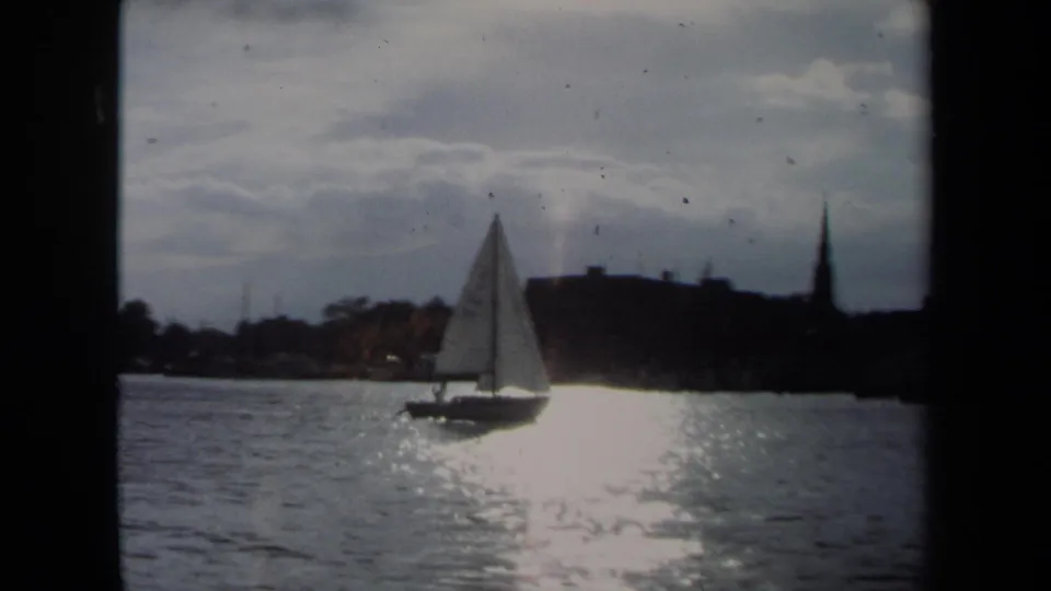 1970 Annapolis harbor view with a white sailboat moving across reflective water and a steeple in the background.