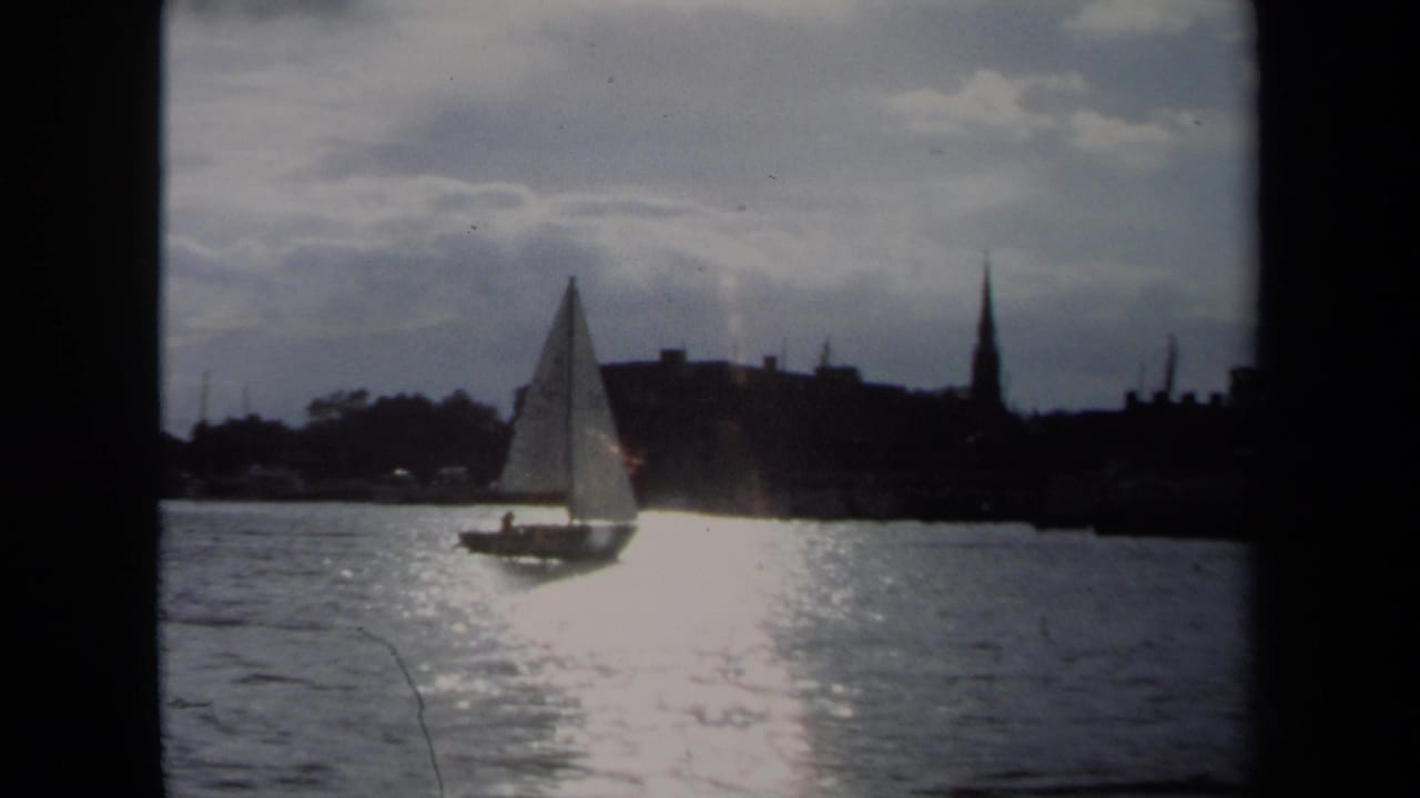A small sailboat continues through Annapolis harbor water under cloudy sky, with bright sun reflection below the sail.