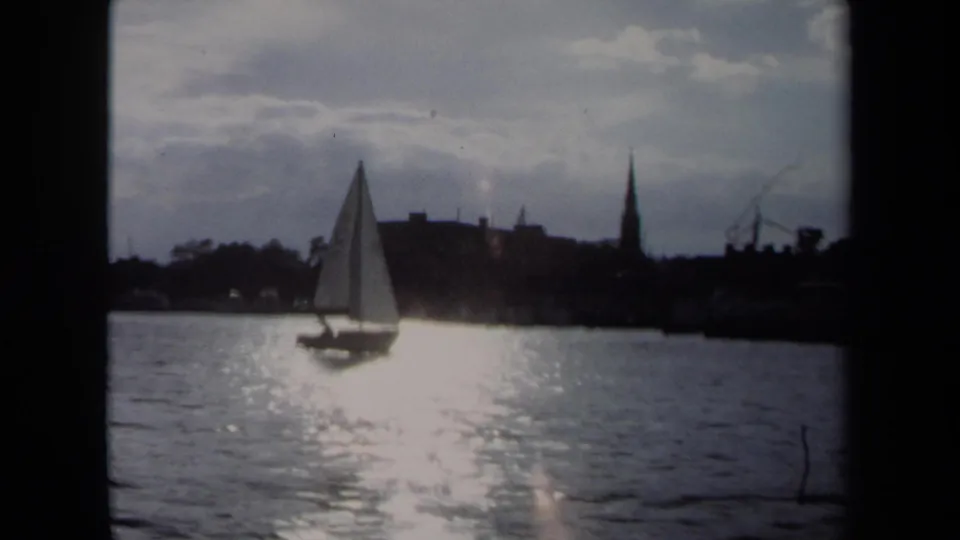 1970 Annapolis sailing view with the boat left of center, sunlit water in front, and a dark waterfront skyline beyond.