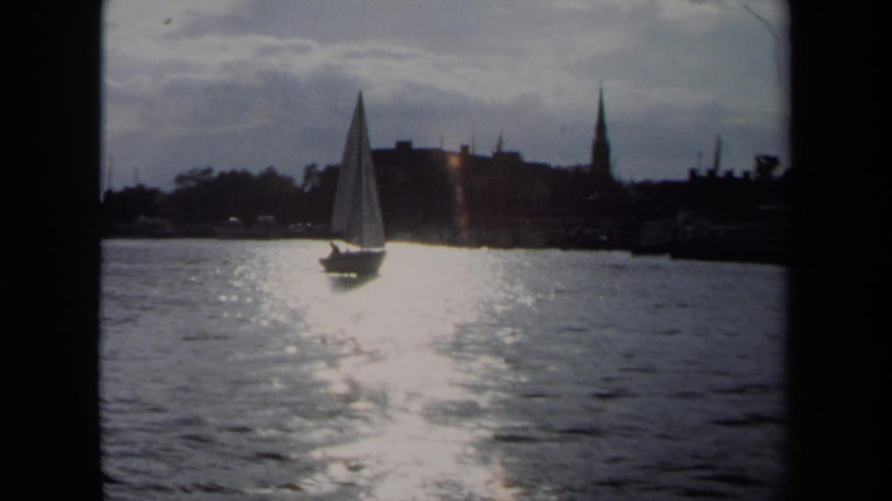 1970 Annapolis 8mm frame showing a sailboat on sunlit water before a silhouetted shoreline with tall steeples.