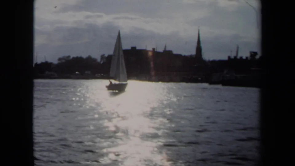 1970 Annapolis 8mm frame showing a sailboat on sunlit water before a silhouetted shoreline with tall steeples.