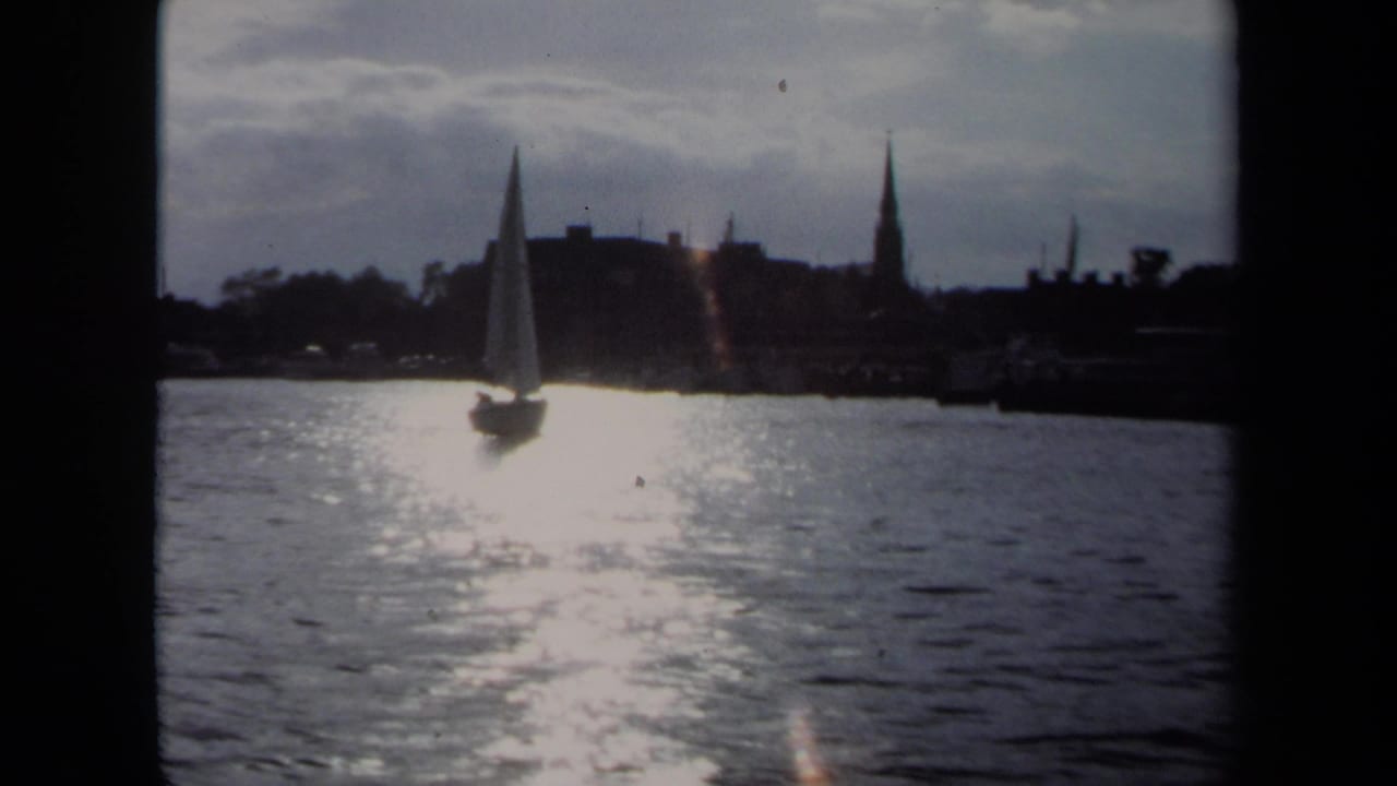 Sailboat on shimmering water with a silhouetted shoreline and steeple behind it.