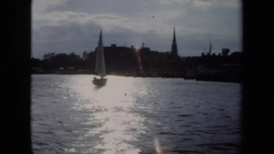1970 Annapolis home-movie frame of a sailboat passing through glare on the water below a dark skyline and steeple.