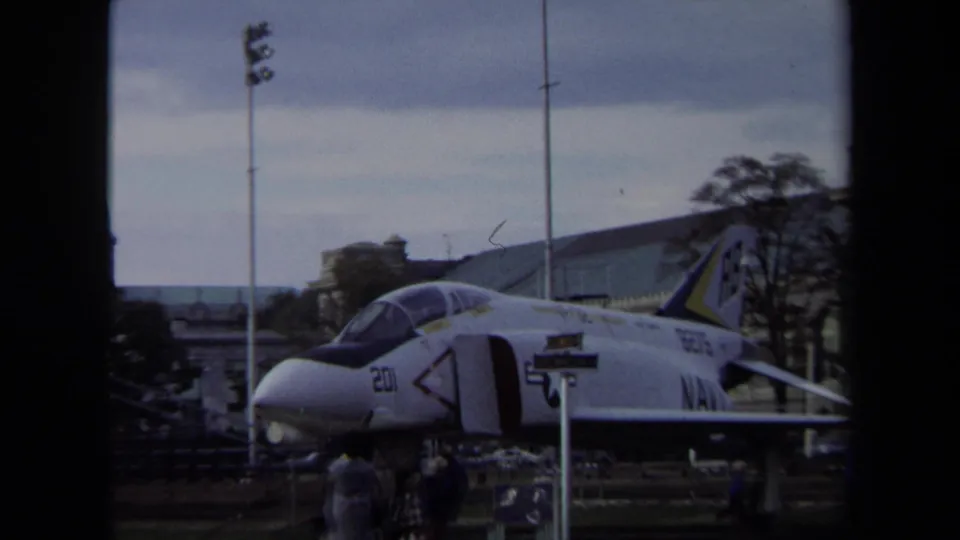 1970 Annapolis 8mm frame showing a U.S. Navy jet display with markings, small visitors, poles, trees, and buildings behind it.