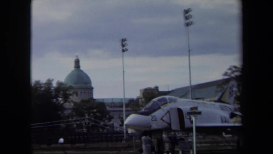1970 Annapolis home-movie frame of a Navy jet display with visible markings, visitors, a green-domed building, poles, and trees.