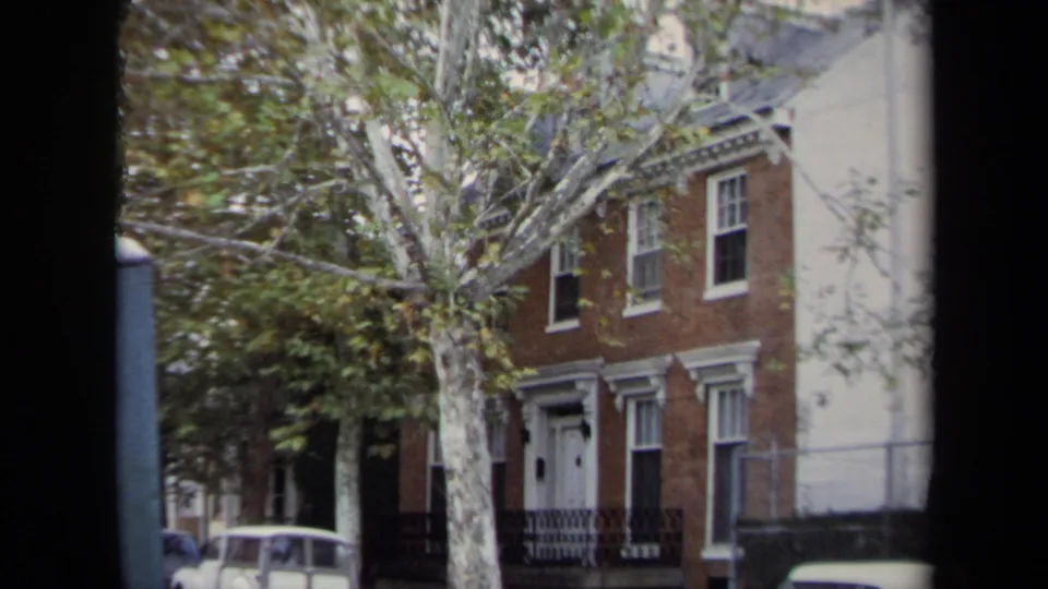 1970 Annapolis home-movie frame of a brick residential facade, street tree, and parked car at curbside.